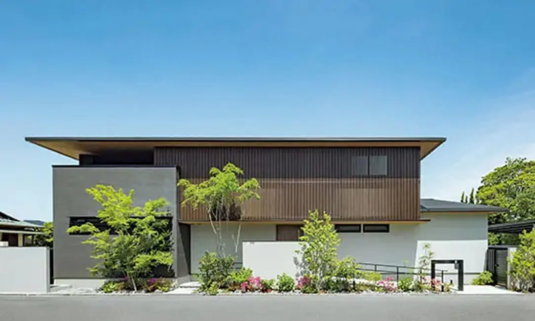 軒の深い屋根と木製ルーバーが特徴の現代的な二階建て住宅。正面には植栽が整えられ、ナチュラルとモダンが融合した外観。