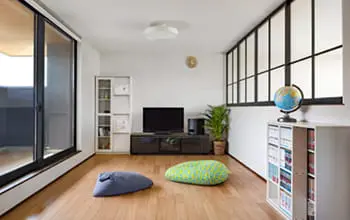 フローリングが敷かれた明るいリビング。テレビと本棚が置かれ、座布団クッションが床に並ぶカジュアルで落ち着いた空間。